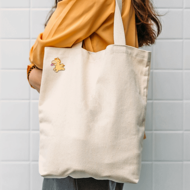 Gif zooming in and out on a image of the animated Bean Blossom Unihorn Pin Badge on a canvas tote bag that's over the shoulder of a girl with a yellow top on