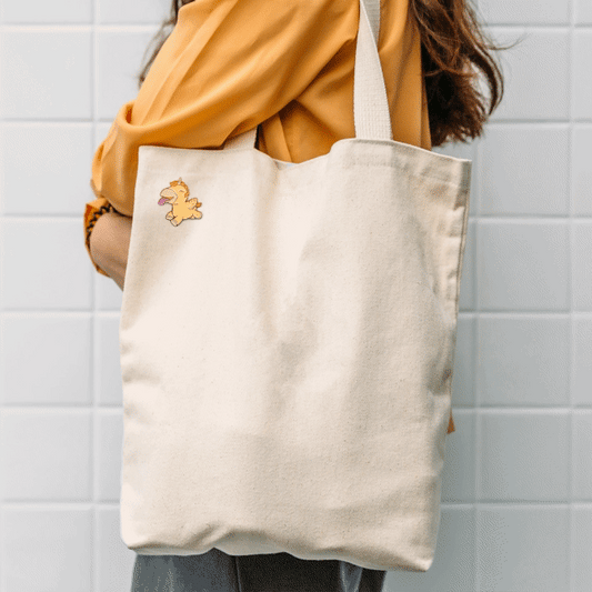Gif zooming in and out on a image of the animated Bean Blossom Unihorn Pin Badge on a canvas tote bag that's over the shoulder of a girl with a yellow top on